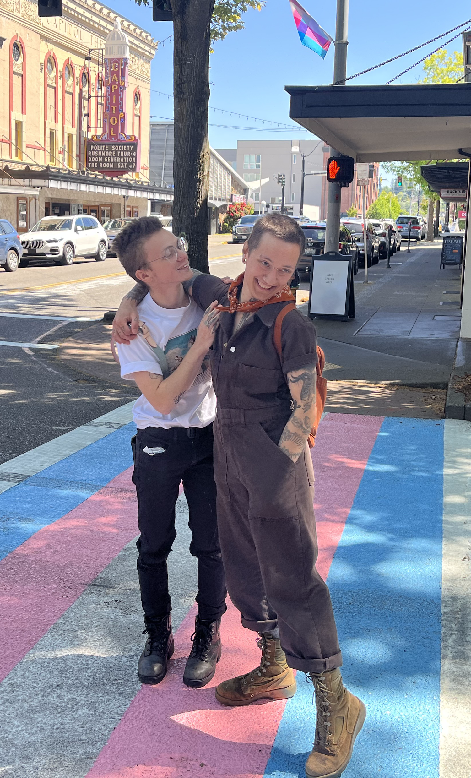 An image of Dimitri and his partner, Ren, with their arms around each other on a sidewalk painted as the trans flag.